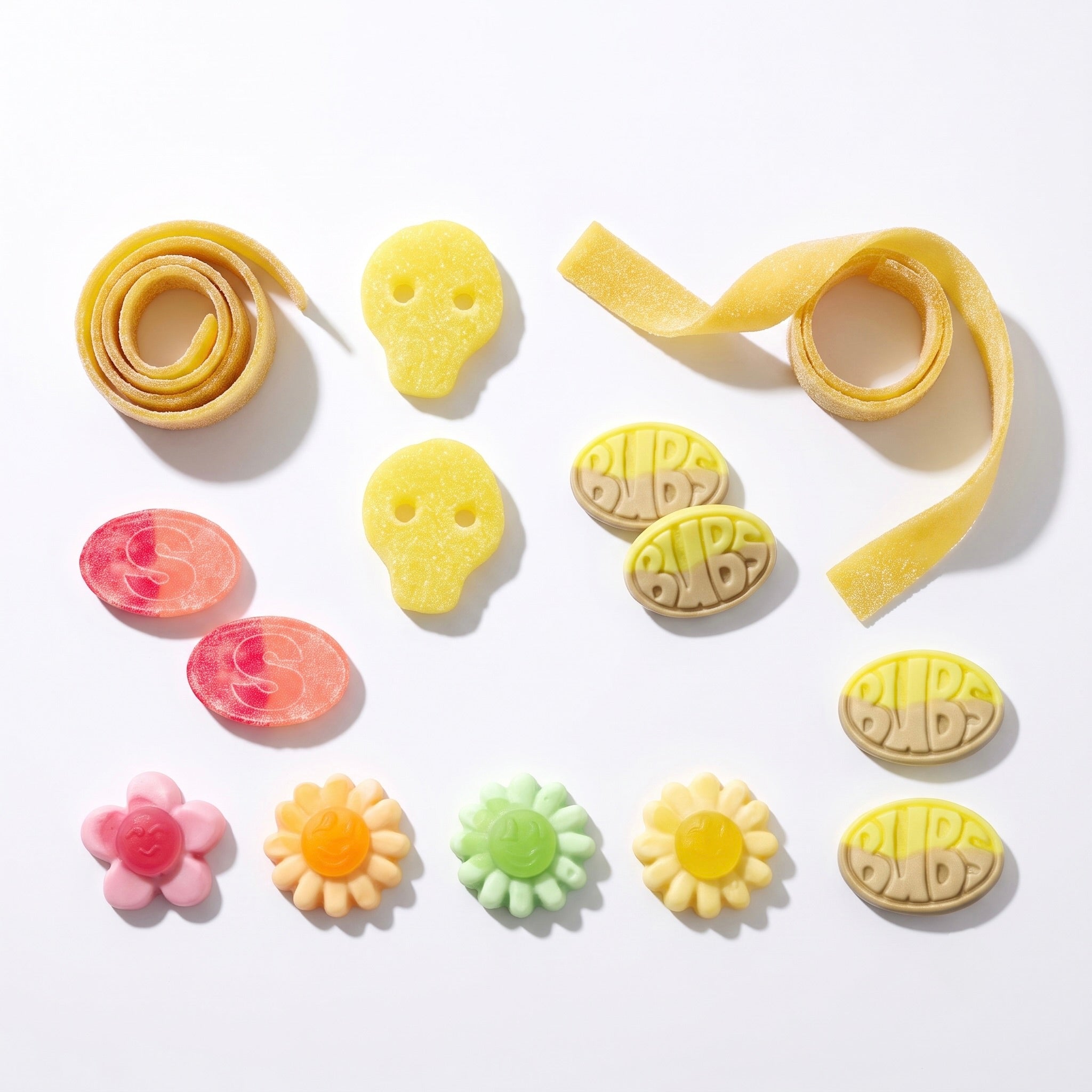 A flat-lay arrangement of colorful pasta-like food pieces on a white background, including yellow mango sour belt, yellow pineapple skull-shaped pieces, pink sour s-marke gummy pieces, yellow BUBS Banana Toffee oval pieces with embossed lettering, a pink flower piece, and yellow, orange, green, and yellow flower pieces.